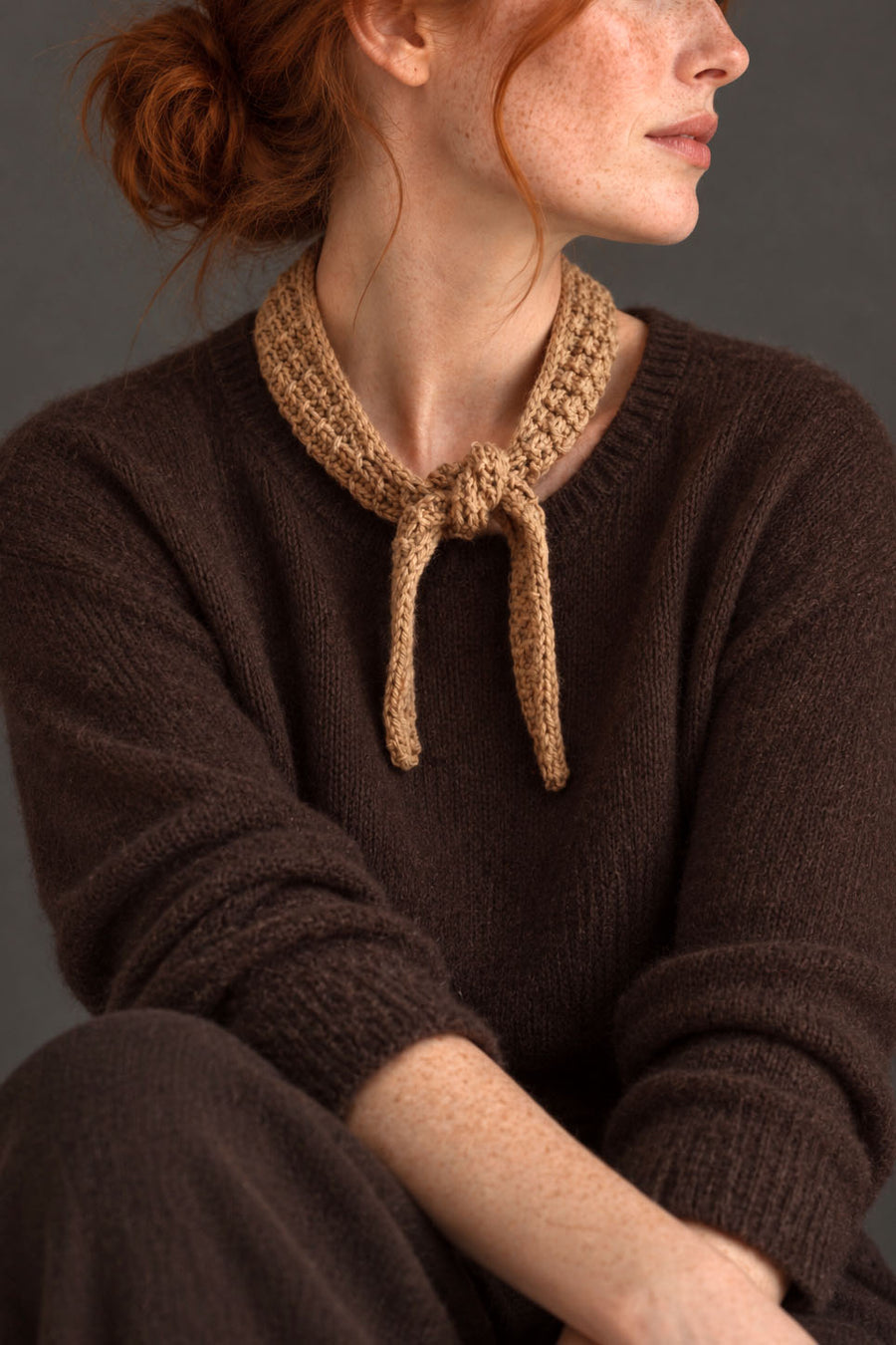 Tan textured skinny scarf tied at the neck over a brown sweater - The Villa Scarf Knitting Pattern by Darling Jadore